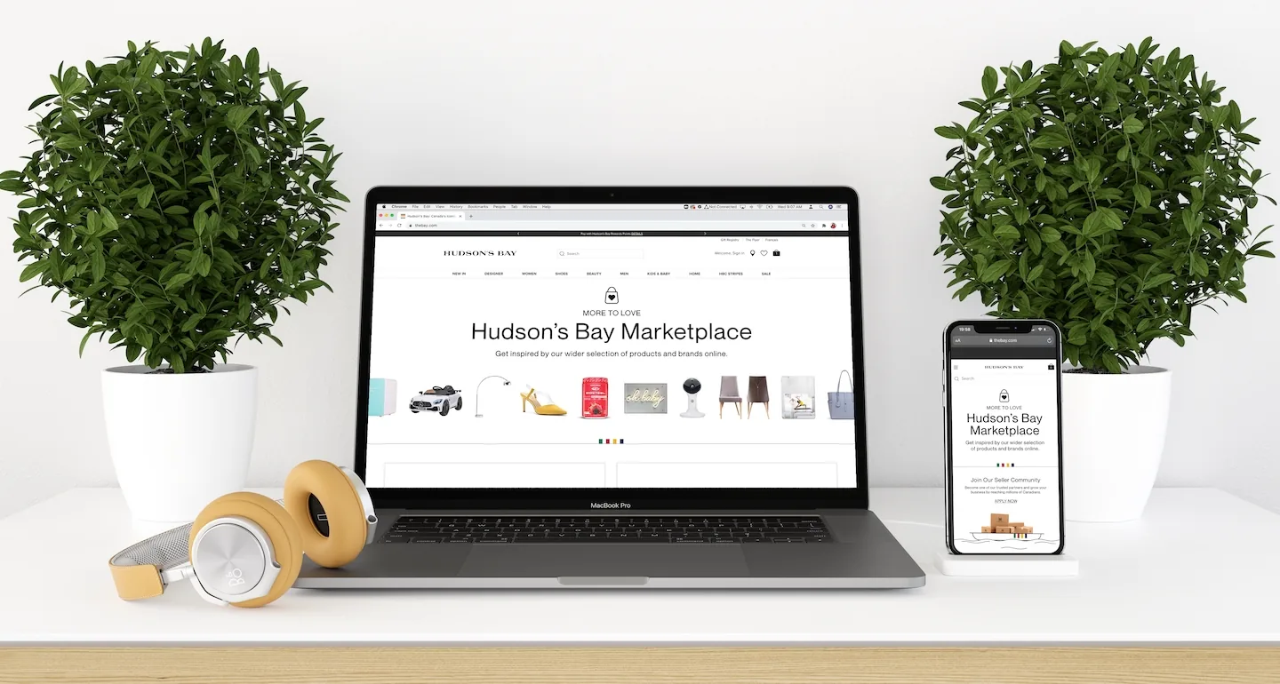 MacBook Pro and iPhone on a desk displaying the Hudson's Bay Marketplace homepage ('More to Love — get inspired by our wider selection of products and brands online'), flanked by potted plants and over-ear headphones