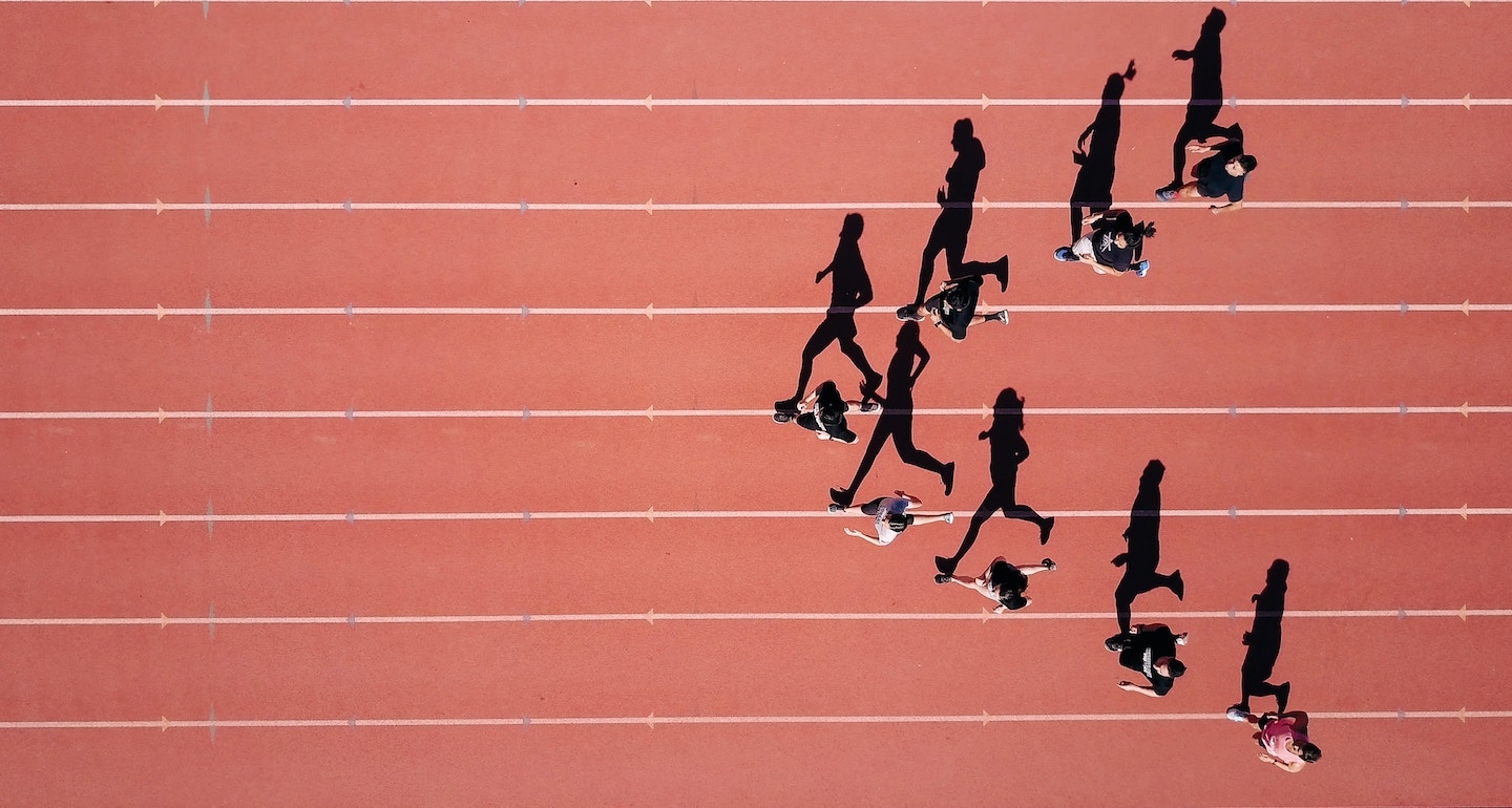 An aerial, birds-eye view of a red running track with several runners casting long, dramatic shadows across the lanes.